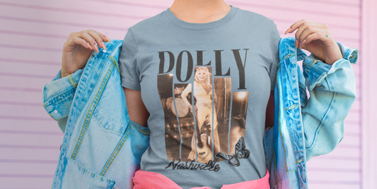 A person wearing a light blue graphic t-shirt with 'DOLLY Nashville' printed on the front, holding light blue denim jackets on both sides against a pink striped background.
