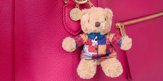 A small plush teddy bear keychain or bag charm hanging from a bright pink textured handbag. The bear is tan and wears a colorful patchwork-style jacket with a blue collar, set against the pink bag surface.