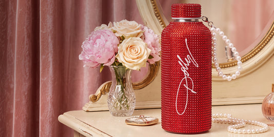 A red rhinestone-covered water bottle with a white script logo that reads "Dolly", shown on a vanity table beside pink flowers in a vase, pearls, and a mirror.