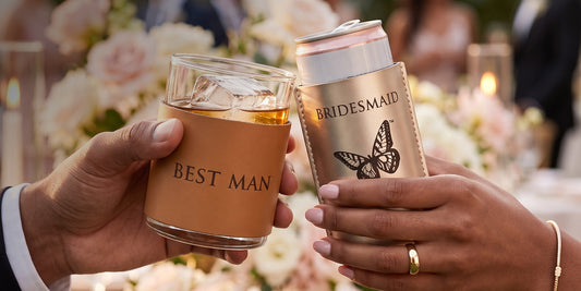 Two hands clinking cocktail glasses in a celebratory toast. One glass has a tan sleeve printed with 'BEST MAN' and the other has a metallic champagne sleeve printed with 'BRIDESMAID' and a butterfly graphic.