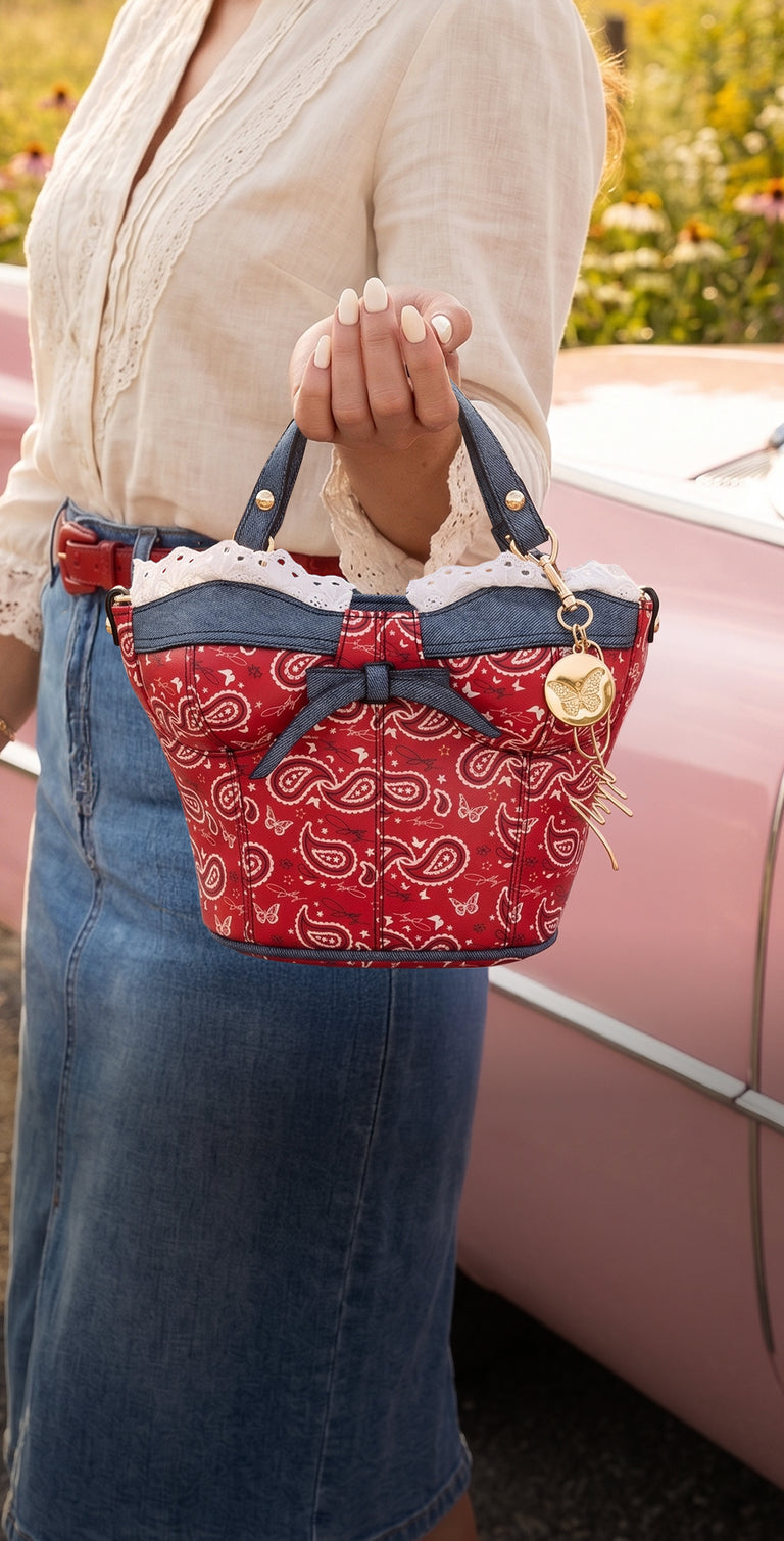 A person holding a red and blue patterned handbag with a bow and gold charm, standing next to a pink car outdoors.