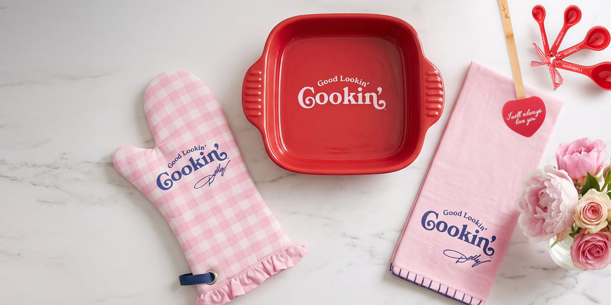 A coordinated kitchen textile set arranged on a light marble surface, including a red rectangular baking pan, a pink gingham oven mitt, a pink towel, and red measuring spoons, all printed with the visible brand name 'Good Lookin' Cookin''.