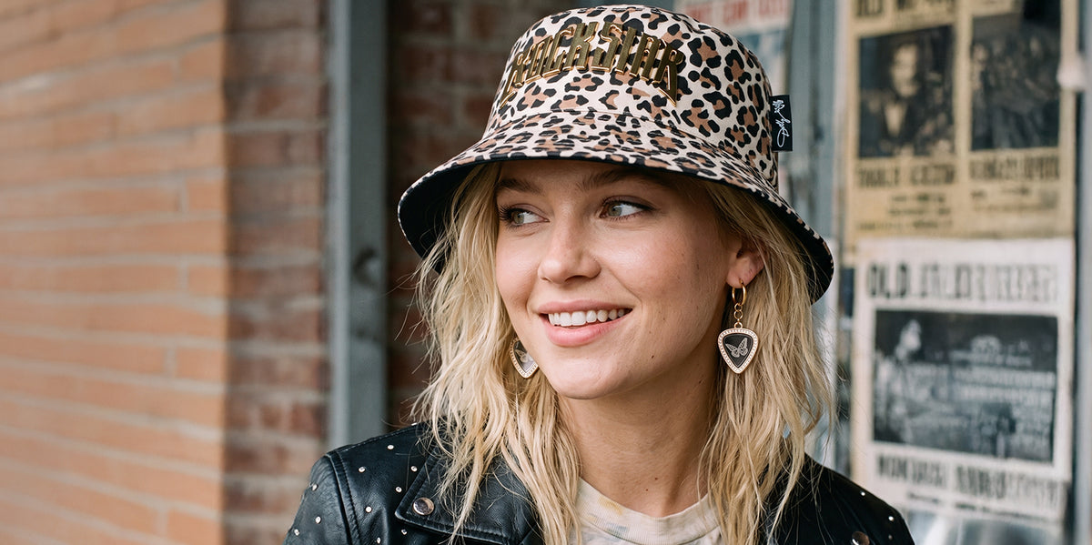 Smiling blonde woman wearing a leopard-print bucket hat with gold 'ROCKSTAR' lettering, guitar-pick butterfly earrings, and a studded black leather jacket, standing in front of a brick wall with vintage concert posters.