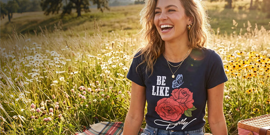 A woman laughing outdoors in a sunlit field of wildflowers, wearing a navy graphic T-shirt with the words “BE LIKE Dolly” and a large red rose print.